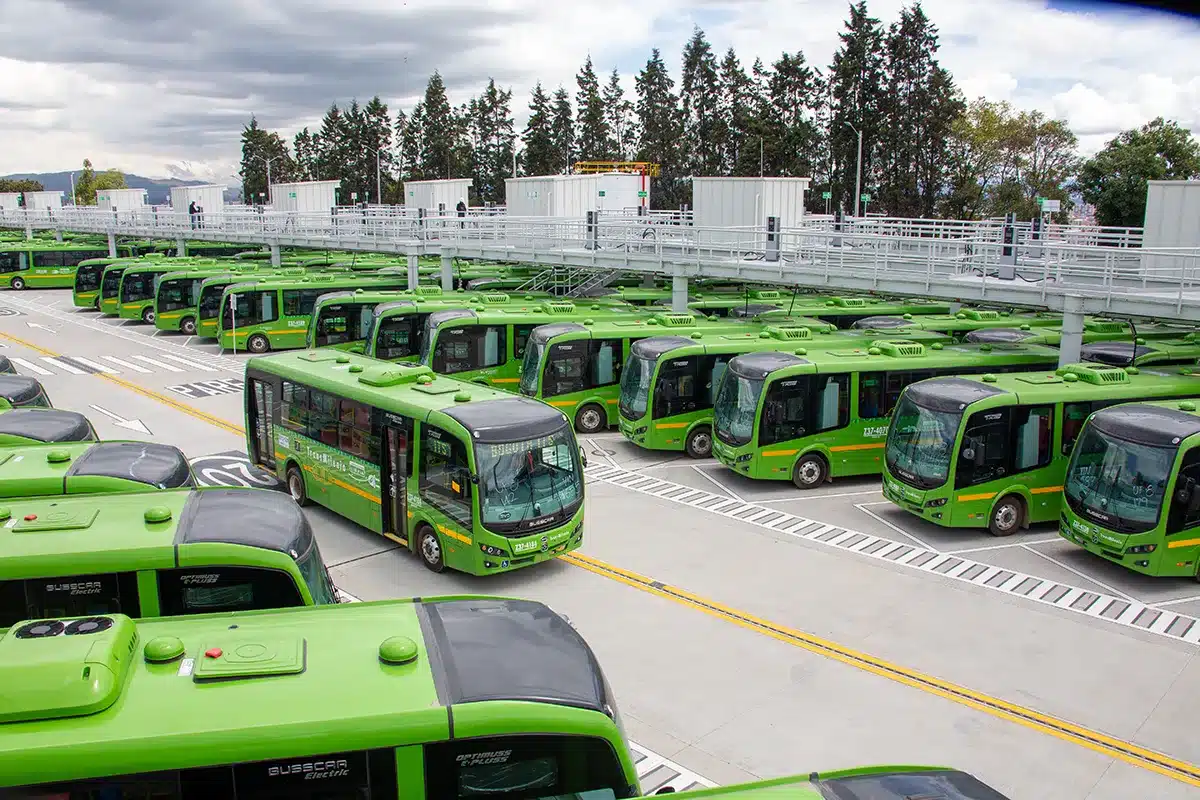 A large fleet of green electric buses at their Bogotá depot under their charging infrastructure, the result of a partnership between VG Mobility and TransMilenio, with one bus leaving the depot