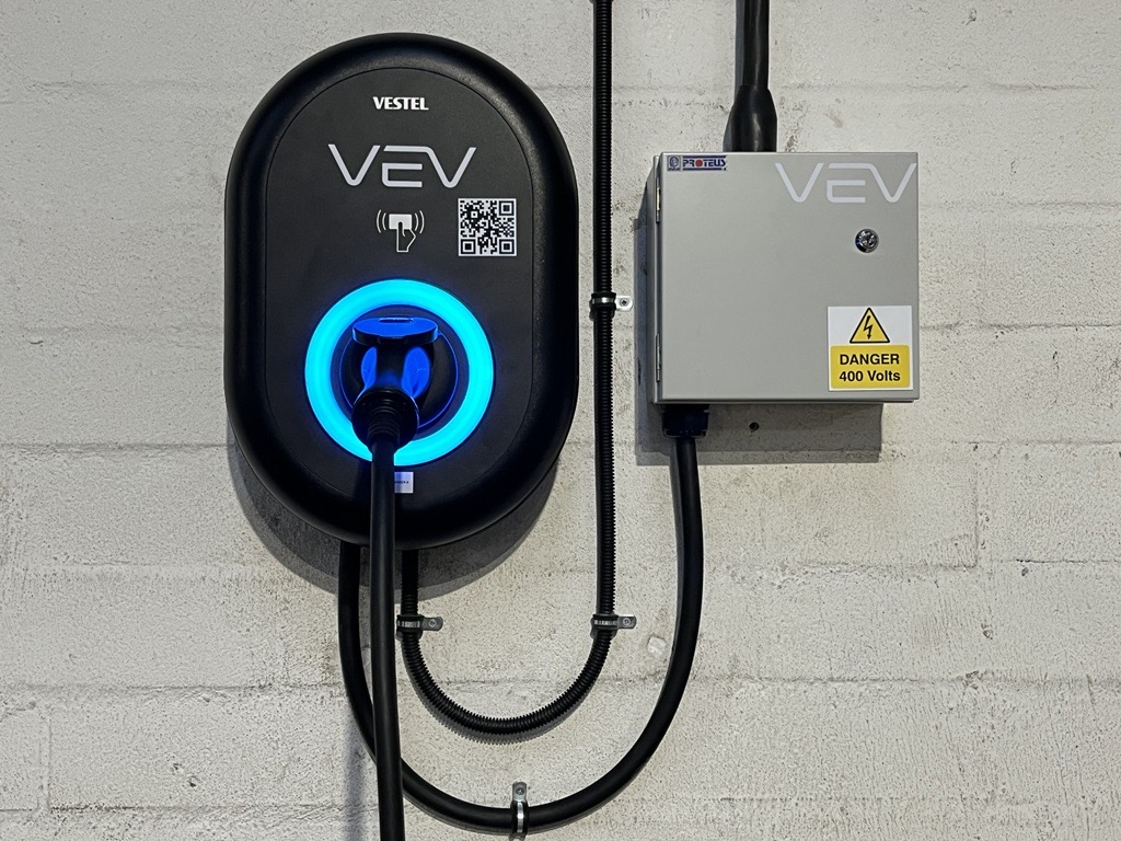 Black electric vehicle charge point with white VEV branding, next to a VEV branded grey inverter box on a white/grey wall